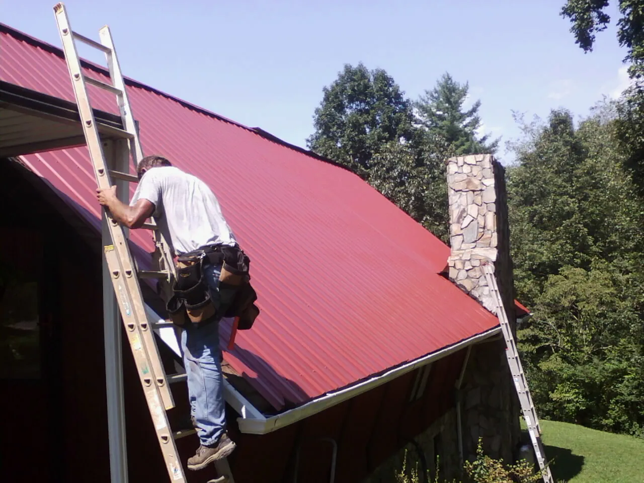 Roofer on a ladder beside a red metal roof and stone chimney — Soffit & Fascia Repair by Roofing Brothers in Riceville
